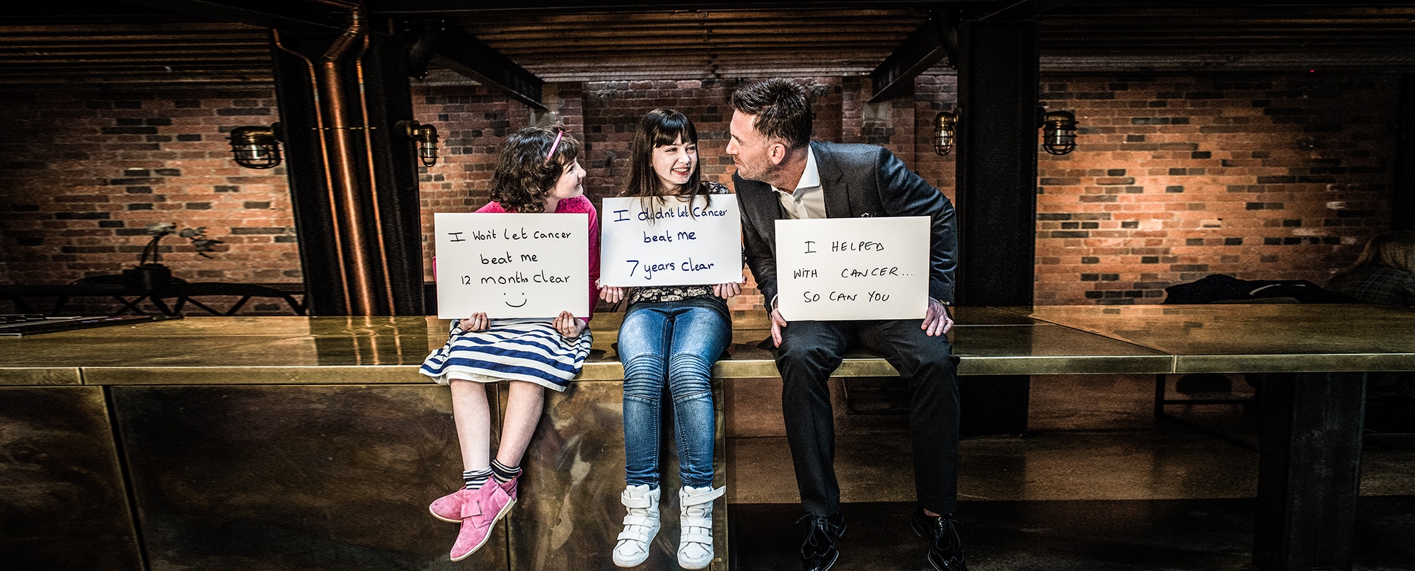 Two young clients and an Optima Hair team member holding handwritten signs about beating cancer